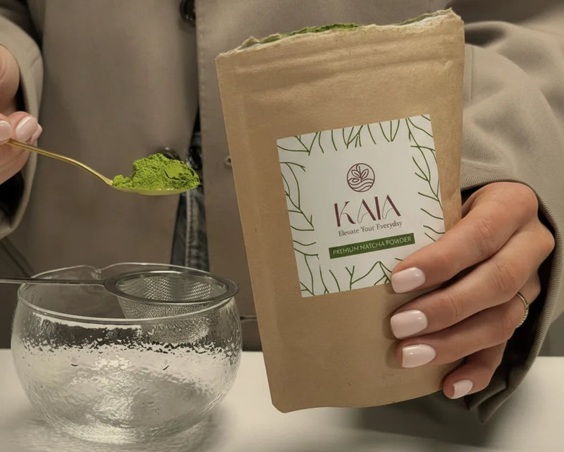 Hand scooping KAIA Origin matcha powder with a spoon, holding a kraft pouch and glass bowl with sieve